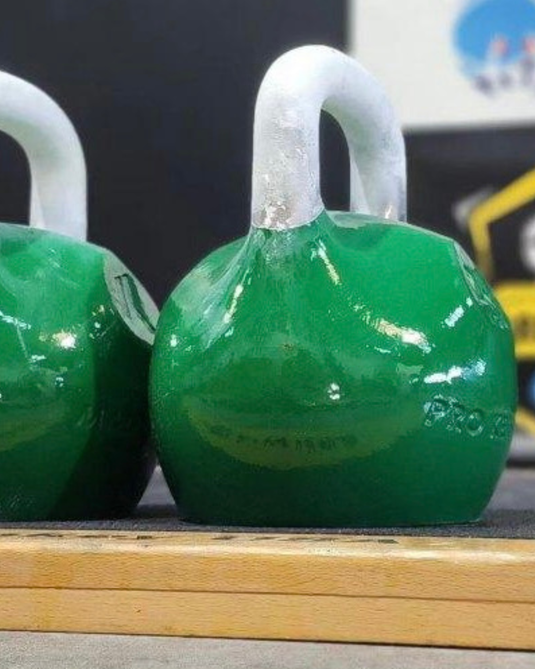 A close-up of a green kettlebell with a white chalked handle resting on a wooden surface, with another kettlebell partially visible on the left.